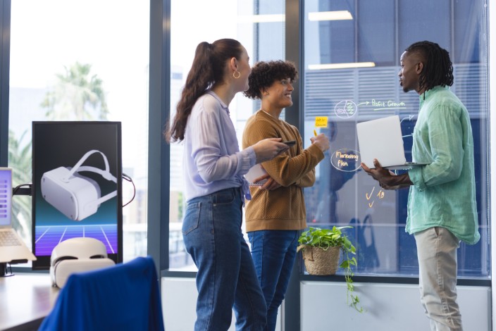 a diverse group of young colleagues discussing in office, holding papers and laptop. the african american man has short hair, caucasian woman wears a purple top, and the biracial man holds the laptop, unaltered