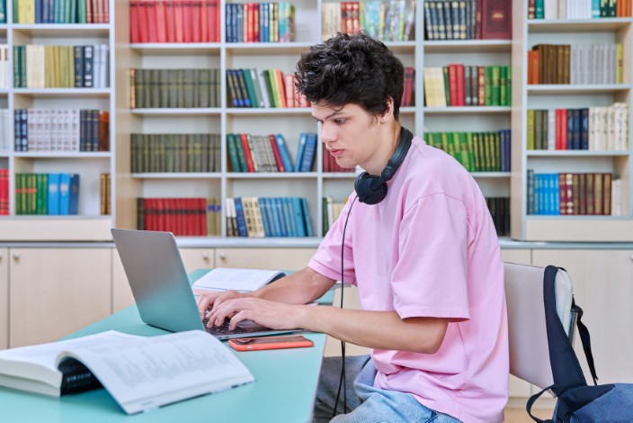 young male student sitting with laptop with books in college library