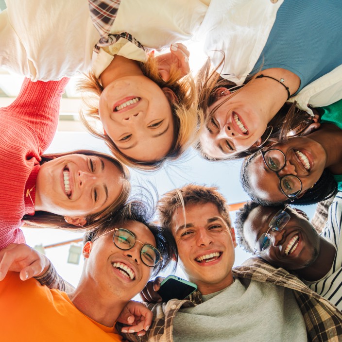 low angle view of a group of multiracial friends standing on a circle, smiling and embracing together. young teenagets laughing and looking at camera. team of confident people on a coaching meeting